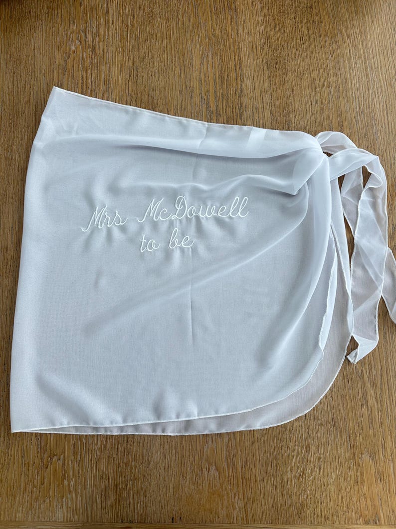 May include: A white wrap skirt made of a sheer, lightweight fabric featuring the embroidered text "Mrs. McDowell to be" in white thread. The garment includes a side tie closure with long fabric straps, creating a layered, flowing silhouette.