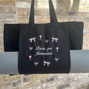 May include: Black canvas tote bag with a yoga mat strapped to the bottom. The bag has pink bows and flowers embroidered on it, and the text "Lava yoo Jennerinio" is embroidered in white.