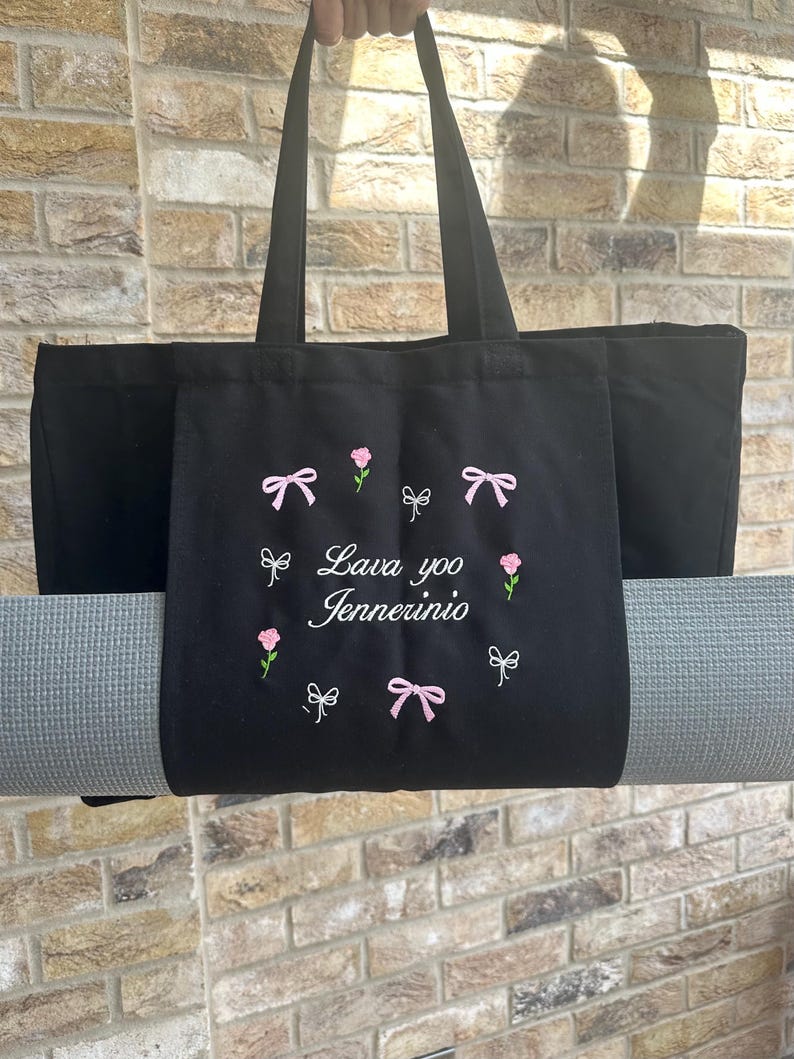 May include: Black canvas tote bag with a yoga mat strapped to the bottom. The bag has pink bows and flowers embroidered on it, and the text "Lava yoo Jennerinio" is embroidered in white.