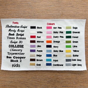 May include: A white fabric swatch featuring an embroidered chart of font styles and color options. The text lists fonts like Ballentines Script and College, alongside color swatches labeled Black, White, Red, Maroon, Lilac, Purple, Beige, Brown, Gold, Hot Pink, Baby Pink, Dusty Pink, Orange, Navy, Baby Blue, Blue, Aqua, Cornflower, Sage, Emerald, Forest, Green, Lime, Yellow, Butter, Dark Grey, and Light Grey.