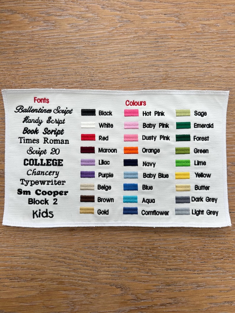May include: A white fabric swatch featuring an embroidered chart of font styles and color options. The text lists fonts like Ballentines Script and College, alongside color swatches labeled Black, White, Red, Maroon, Lilac, Purple, Beige, Brown, Gold, Hot Pink, Baby Pink, Dusty Pink, Orange, Navy, Baby Blue, Blue, Aqua, Cornflower, Sage, Emerald, Forest, Green, Lime, Yellow, Butter, Dark Grey, and Light Grey.