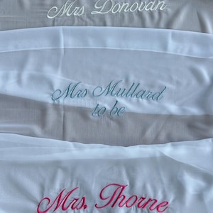 May include: Three white fabric sashes featuring embroidered text in cursive script. The top sash reads Mrs. Donovan in white, the middle sash reads Mrs. Mullard to be in light blue, and the bottom sash reads Mrs. Thorne in bright pink embroidery.