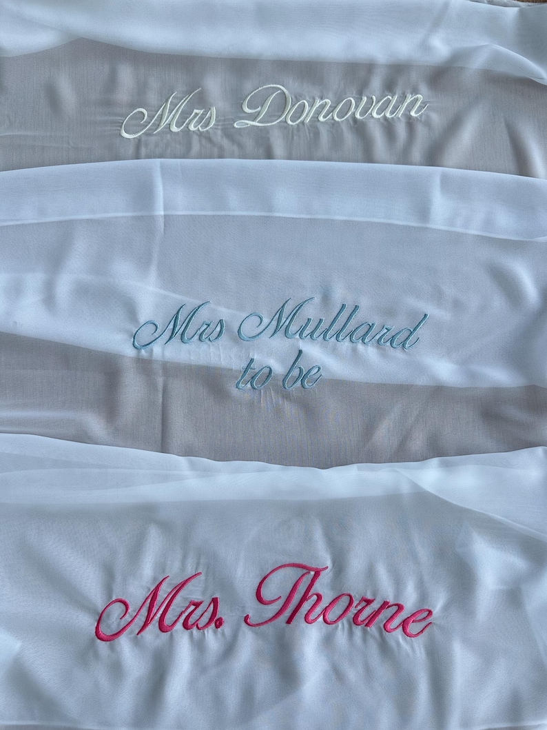 May include: Three white fabric sashes featuring embroidered text in cursive script. The top sash reads Mrs. Donovan in white, the middle sash reads Mrs. Mullard to be in light blue, and the bottom sash reads Mrs. Thorne in bright pink embroidery.