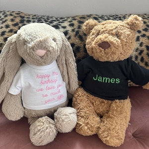 May include: Two soft toys: a beige bunny wearing a white t-shirt with pink text saying "happy 21st birthday! we love you so much - your sis" and a brown teddy bear in a black t-shirt with "James" in green.