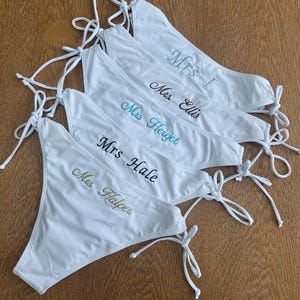 May include: A stack of five white bikini bottoms with side ties. Each piece features custom embroidery in different colors: Mrs. I in light blue, Mrs. Ellis in dark blue, Mrs. Herget in teal, Mrs. Hale in black, and Mrs. Halper in gold thread.