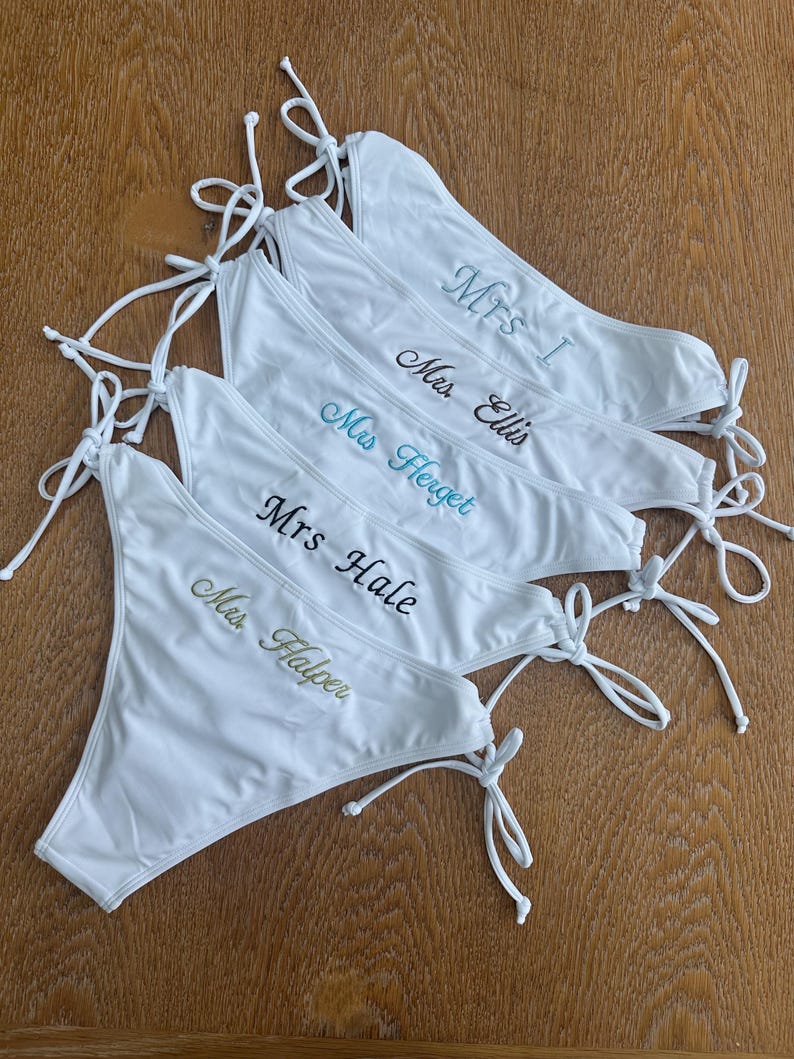 May include: A stack of five white bikini bottoms with side ties. Each piece features custom embroidery in different colors: Mrs. I in light blue, Mrs. Ellis in dark blue, Mrs. Herget in teal, Mrs. Hale in black, and Mrs. Halper in gold thread.