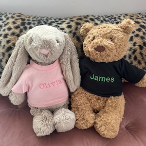 May include: Two soft toys: a bunny and a teddy bear. The bunny is light grey and wears a pink top embroidered with "Olivia." The teddy bear is brown and wears a black top embroidered with "James."