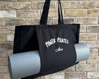 May include: Black tote bag with a gray yoga mat. The bag has the words "POWER PILATES" and the name "Alex" printed in white. The bag is held up against a brick wall.