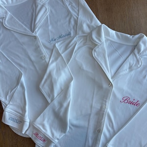 May include: Two white pajama tops with button-down fronts and notched collars. One top has "Mrs. Mitchell" embroidered in blue, the other has "Bride" in pink. Dates are embroidered on the cuffs.