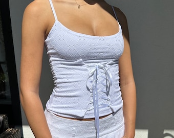 May include: White lace corset top with a white ribbon tie in the front. The top is sleeveless and has a scoop neck.