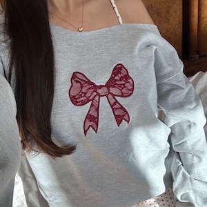 May include: A gray off-the-shoulder sweatshirt with a red lace bow embroidered on the front. The sweatshirt is paired with white shorts with a red heart pattern.