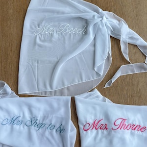May include: Three white sheer wrap skirts featuring embroidered text. One skirt displays 'Mrs. Beech' in white thread, another shows 'Mrs. Shep to be' in blue thread, and the third features 'Mrs. Thorne' in pink thread. Each skirt has a tie closure.