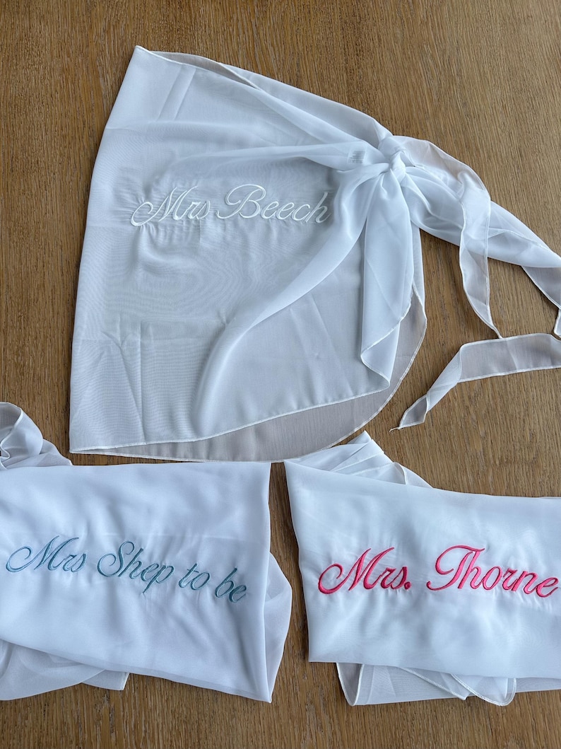 May include: Three white sheer wrap skirts featuring embroidered text. One skirt displays 'Mrs. Beech' in white thread, another shows 'Mrs. Shep to be' in blue thread, and the third features 'Mrs. Thorne' in pink thread. Each skirt has a tie closure.