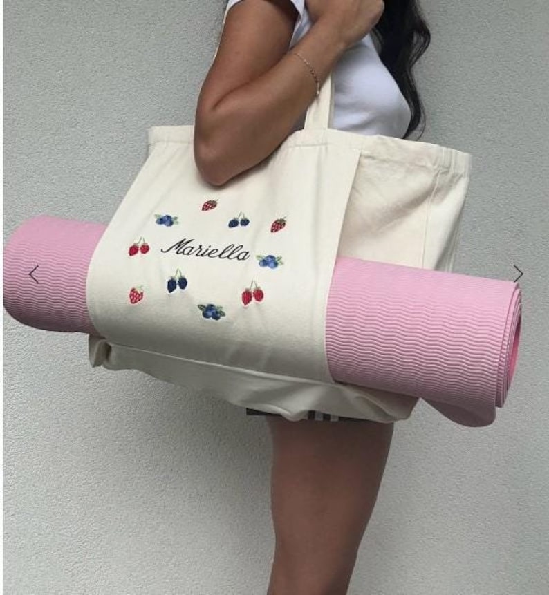 May include: A white canvas tote bag with the name "Mariella" embroidered in black. The bag has a pink yoga mat sticking out of it and is decorated with embroidered red strawberries and blue blueberries.