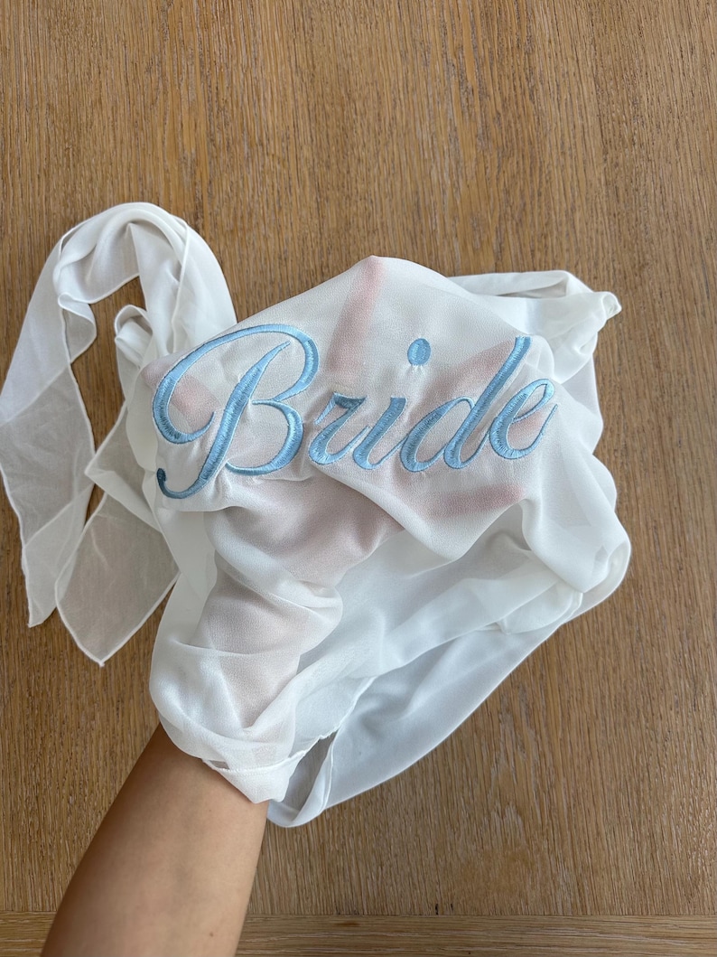 May include: A sheer white fabric garment featuring the word Bride embroidered in light blue cursive script. The lightweight, semi-transparent material is draped, revealing the texture of the fabric and the detailed blue stitching of the lettering.