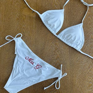 May include: A white two-piece bikini set featuring a triangle top with thin tie straps and matching bikini bottoms with side ties. The bikini bottoms are embroidered with the red cursive text "Mrs. C" on the front left side of the fabric.