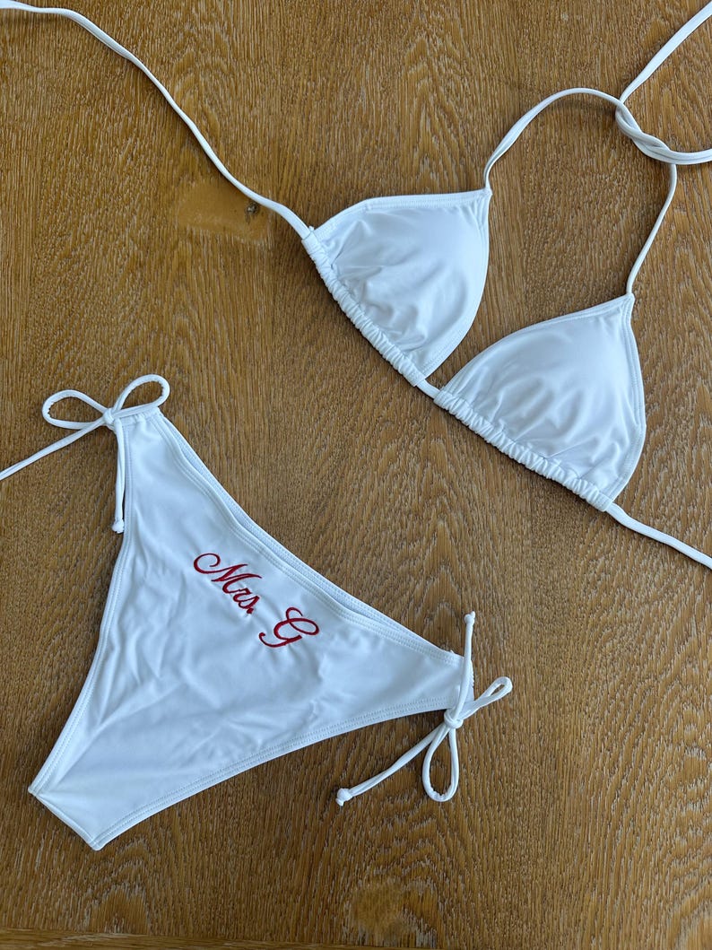 May include: A white two-piece bikini set featuring a triangle top with thin tie straps and matching bikini bottoms with side ties. The bikini bottoms are embroidered with the red cursive text "Mrs. C" on the front left side of the fabric.