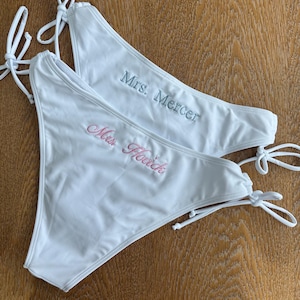 May include: Two white bikini bottoms with side ties. One bottom features the embroidered text "Mrs. Mercer" in light blue, while the other features the embroidered text "Mrs. Hovick" in light pink. Both garments are plain white with matching white ties.