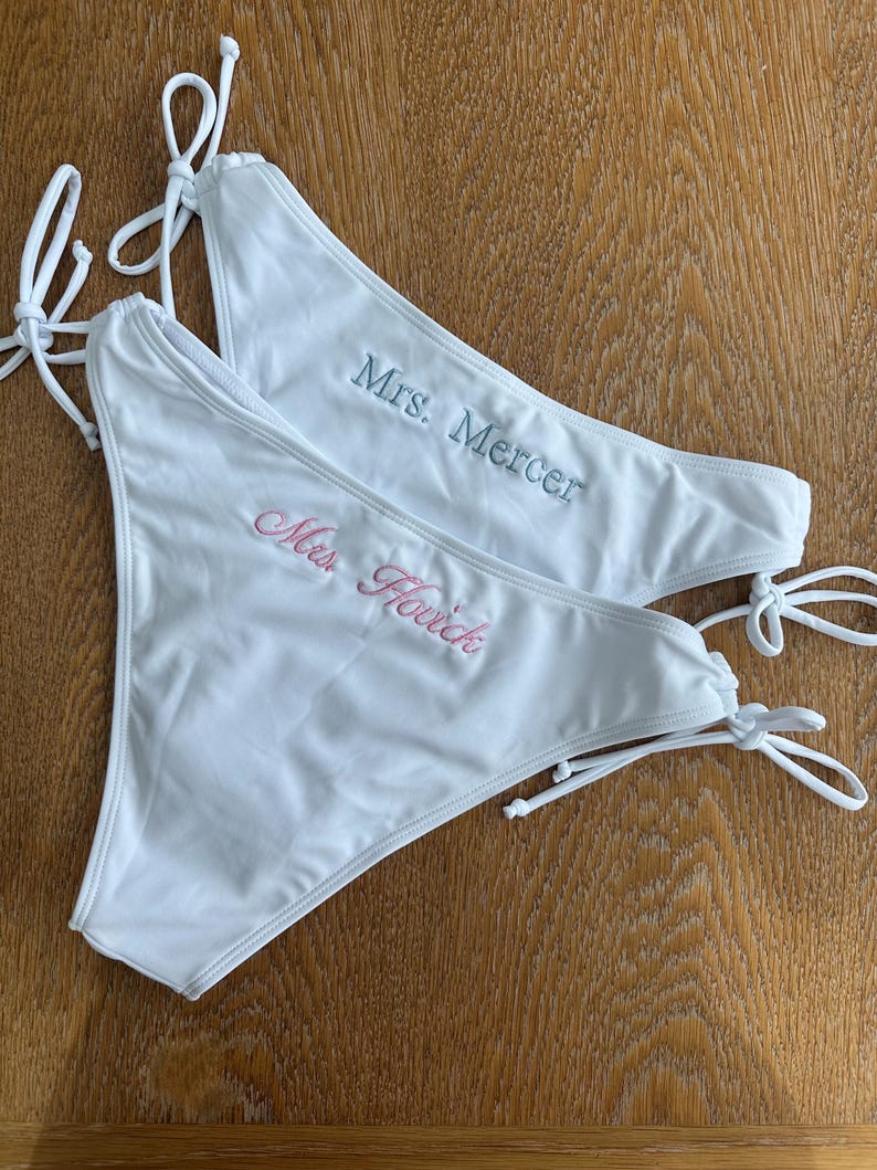 May include: Two white bikini bottoms with side ties. One bottom features the embroidered text "Mrs. Mercer" in light blue, while the other features the embroidered text "Mrs. Hovick" in light pink. Both garments are plain white with matching white ties.