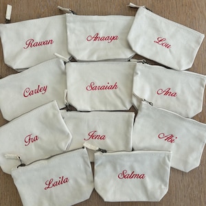 Peut inclure: Une collection de dix pochettes zippées en toile blanc cassé, chacune portant un prénom brodé en écriture rouge. Les prénoms incluent Rawan, Anaaya, Lou, Carley, Saraiah, Ana, Fra, Jena, Abi, Laila et Salma, disposés en une grille.