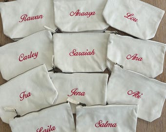 May include: A collection of ten off-white canvas zipper pouches, each featuring a unique name embroidered in red cursive script. The names include Rawan, Anaaya, Lou, Carley, Saraiah, Ana, Fra, Jena, Abi, Laila, and Salma, all with metal zippers.
