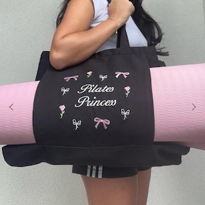 May include: Black tote bag with a pink yoga mat inside. The bag has white embroidery that says "Pilates Princess" with pink bows and flowers.
