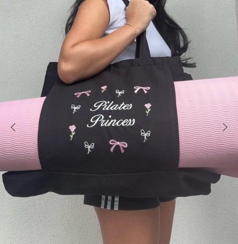 May include: Black tote bag with a pink yoga mat inside. The bag has white embroidery that says "Pilates Princess" with pink bows and flowers.