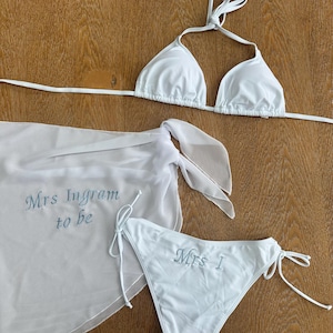 May include: A white bikini set consisting of a triangle top and side-tie bottoms, accompanied by a matching sheer sarong. The sarong features the embroidered text "Mrs Ingram to be" in blue, while the bikini bottom displays "Mrs I" in blue.