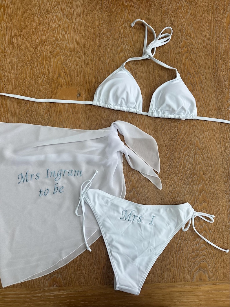 May include: A white bikini set consisting of a triangle top and side-tie bottoms, accompanied by a matching sheer sarong. The sarong features the embroidered text "Mrs Ingram to be" in blue, while the bikini bottom displays "Mrs I" in blue.