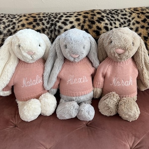 May include: Three soft toy bunny rabbits in white, grey, and beige, each wearing a pink jumper with a name embroidered on it. The bunnies are on a pink surface with a leopard print background.