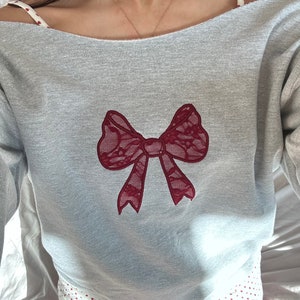 May include: A gray sweatshirt with a red lace bow design. The sweatshirt has a scoop neck and long sleeves.