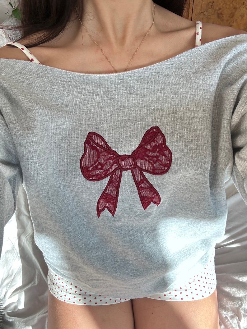 May include: A gray sweatshirt with a red lace bow design. The sweatshirt has a scoop neck and long sleeves.