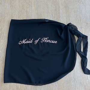 May include: A black wrap skirt made of sheer fabric featuring the text Maid of Honour embroidered in white script lettering. The garment includes a side tie closure with long fabric straps, creating a layered appearance with a curved hemline.