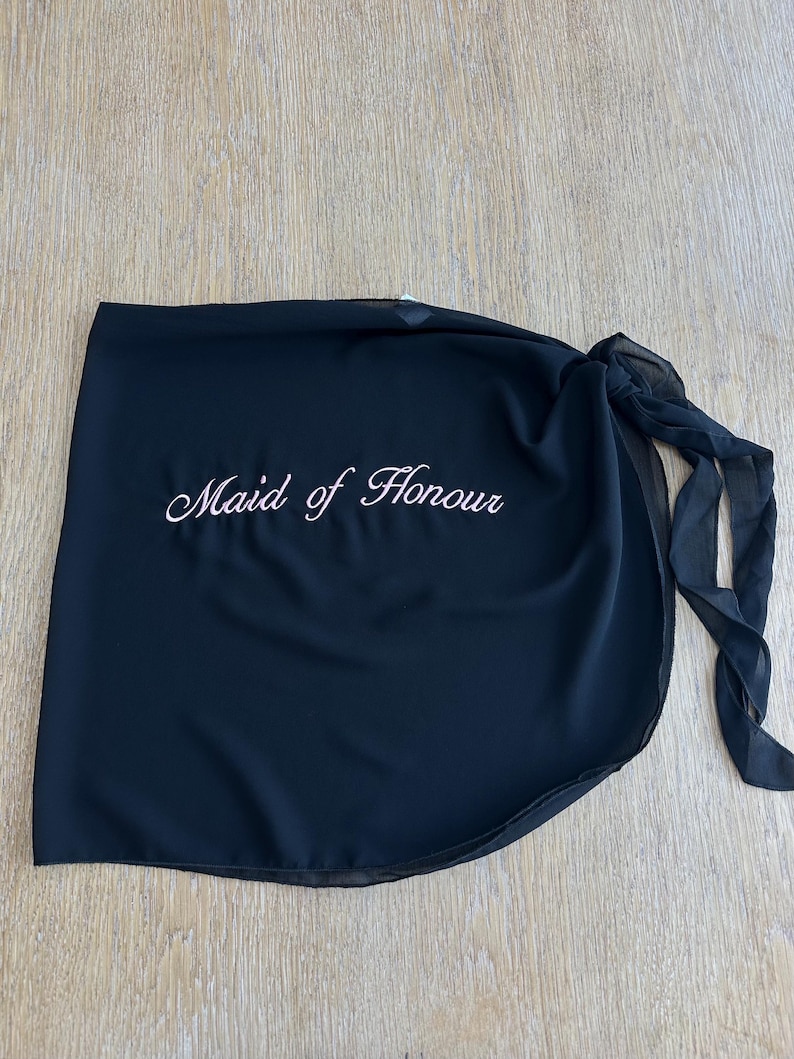 May include: A black wrap skirt made of sheer fabric featuring the text Maid of Honour embroidered in white script lettering. The garment includes a side tie closure with long fabric straps, creating a layered appearance with a curved hemline.