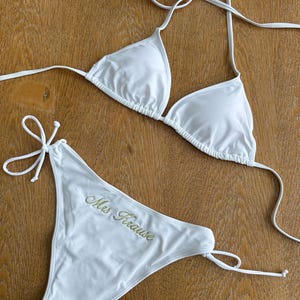 May include: A white two-piece bikini set featuring a triangle top with thin halter straps and matching tie-side bikini bottoms. The front of the bikini bottoms features the embroidered text Mrs Krause in a gold-colored script font.