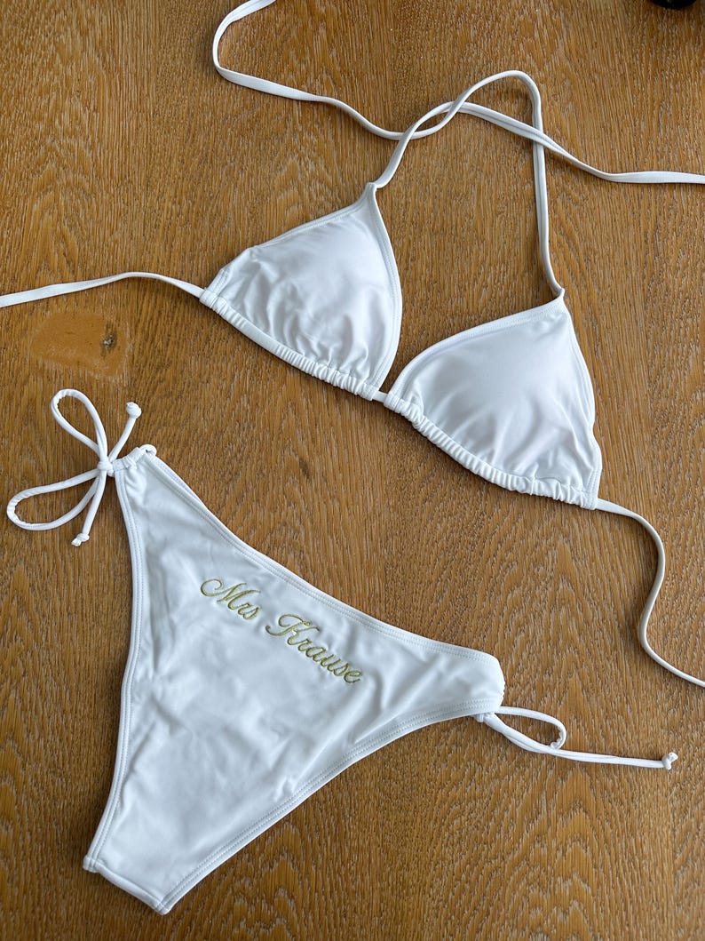 May include: A white two-piece bikini set featuring a triangle top with thin halter straps and matching tie-side bikini bottoms. The front of the bikini bottoms features the embroidered text Mrs Krause in a gold-colored script font.