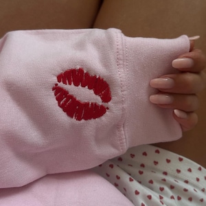 May include: A pale pink sweatshirt sleeve featuring a prominent embroidered red kiss mark. The sleeve rests on a patterned surface. The image also shows a hand with long, polished nails.