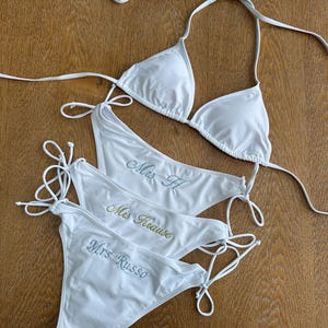 May include: White bikini set with triangle top and string tie bottoms. The bottoms are embroidered with the names "Mrs H", "Mrs Krause", and "Mrs Russo" in different colors. The swimwear is laid out on a wooden surface.