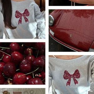 May include: A gray off-the-shoulder sweatshirt with a burgundy lace bow on the front. The sweatshirt is worn by a person with long dark hair. The background is a blurry image of a room.