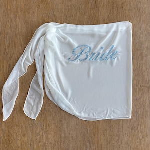 May include: A white sheer wrap skirt featuring the word Bride embroidered in light blue script lettering. The garment has a tie-side closure with long, flowing fabric panels that drape naturally, creating a soft and lightweight appearance.