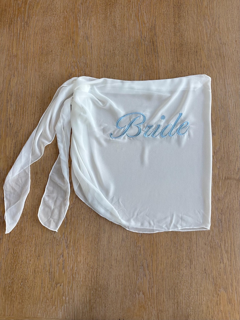 May include: A white sheer wrap skirt featuring the word Bride embroidered in light blue script lettering. The garment has a tie-side closure with long, flowing fabric panels that drape naturally, creating a soft and lightweight appearance.