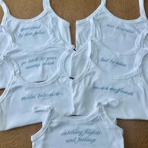 May include: A collection of white vest tops with thin straps, each featuring unique teal embroidered text. The phrases include "sponsored by don julio," "mrs. bhandal," "go back to your swamp, shrek," "bed by nine," "model behaviour," "excuse my french," and "catching flights and feelings."