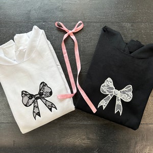 May include: Two hooded sweatshirts, one white and one black, with a white lace bow design on each. A pink ribbon is lying on top of the sweatshirts.