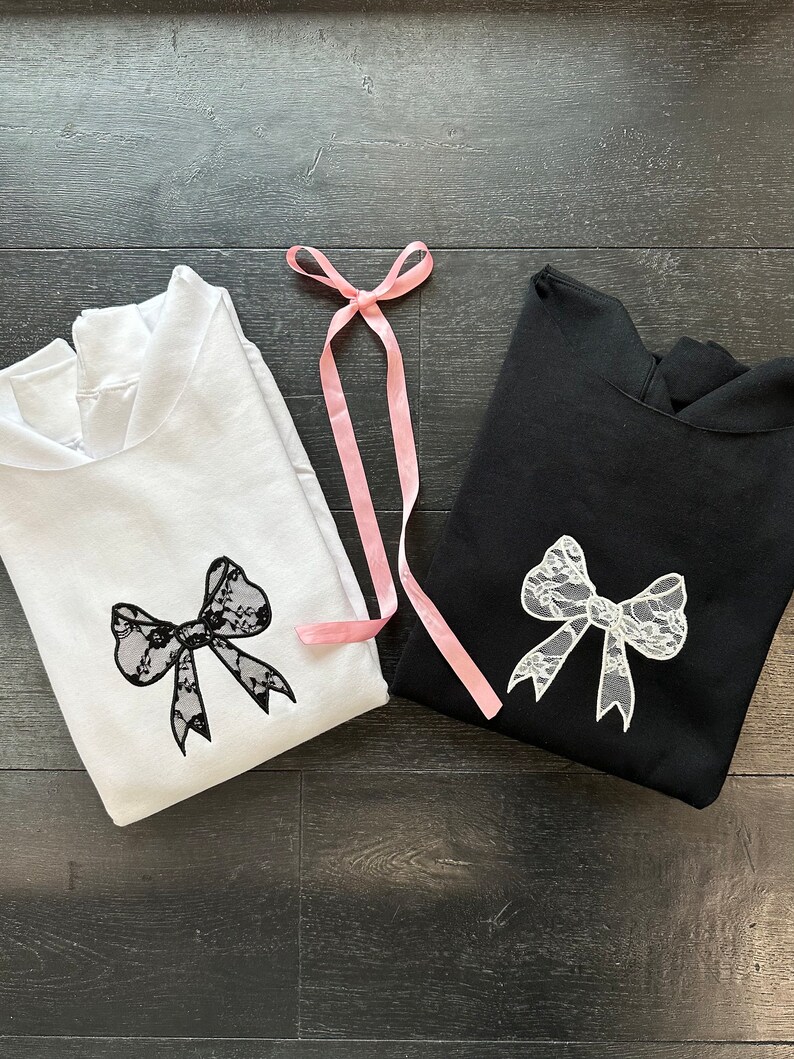 May include: Two hooded sweatshirts, one white and one black, with a white lace bow design on each. A pink ribbon is lying on top of the sweatshirts.