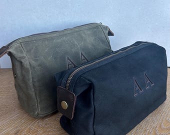 May include: Two rectangular toiletry bags made of canvas, one in olive green and one in black. Each bag features a top zip closure, brown leather side tabs with brass-coloured metal snaps, and the embossed capital letters AA on the front.