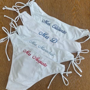 May include: Four white bikini bottoms with side ties, each featuring custom embroidery. The text reads Mrs. Ciliberto in dark blue, Mrs. D in blue, Mrs. Marechal in light blue, and Mrs. Augusto in red script lettering across the front.