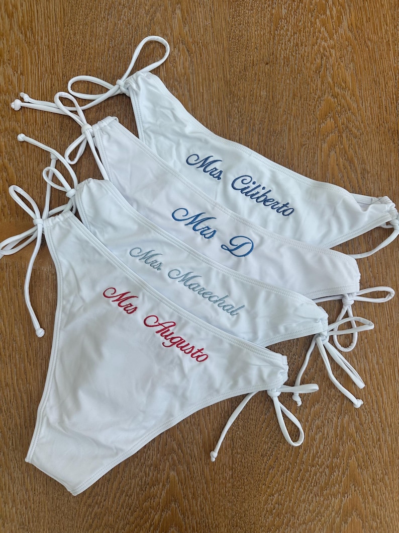 May include: Four white bikini bottoms with side ties, each featuring custom embroidery. The text reads Mrs. Ciliberto in dark blue, Mrs. D in blue, Mrs. Marechal in light blue, and Mrs. Augusto in red script lettering across the front.