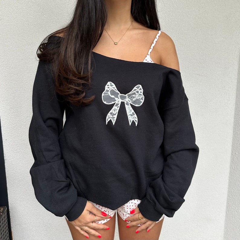 May include: Black off-the-shoulder sweatshirt with a white lace bow design.