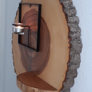 Könnte beinhalten: Ein rustikaler Wandleuchter aus Holz mit natürlicher Kante. Er hat einen schwarzen Metallrahmen, der einen kleinen Kerzenhalter mit einer brennenden Kerze hält. Unter dem Kerzenhalter befindet sich ein kleines Holzregal. Das Holz hat eine hellbraune Farbe.