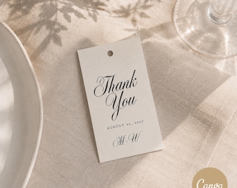 May include: A rectangular off-white paper tag with a hole at the top. The text reads Thank You in a black script font, followed by the date AUGUST 25, 2027 and the initials M.W in a serif font. The tag is positioned on a textured fabric surface.